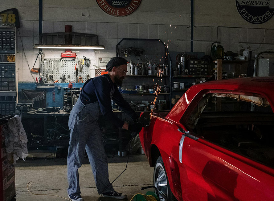 A man repairs a red car inside a garage, surrounded by tools and equipment.
