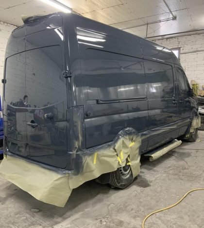 A van is being painted inside a garage, with paint supplies and tools visible in the background.