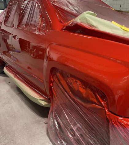 A shiny red pickup truck is partially covered in plastic wrap, indicating it’s in the process of being painted in a workshop.