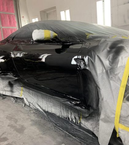 A car is being painted in a garage, with tools and paint supplies visible in the background.