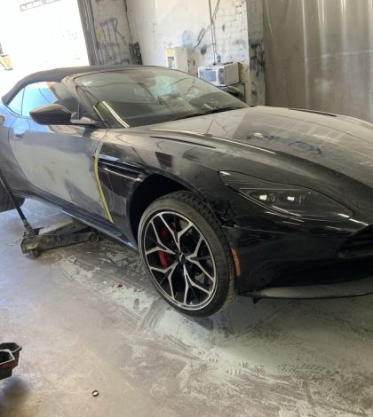 A black Aston Martin car is being painted inside a garage, with tools and paint supplies visible in the background.