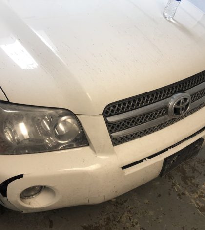 A white Toyota Highlander with a visibly damaged bumper parked on a street.