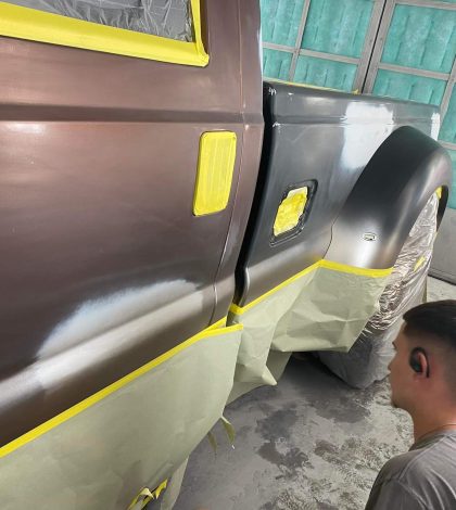 A partial view of a vehicle being prepped for painting, with sections covered in masking tape and a person working nearby.