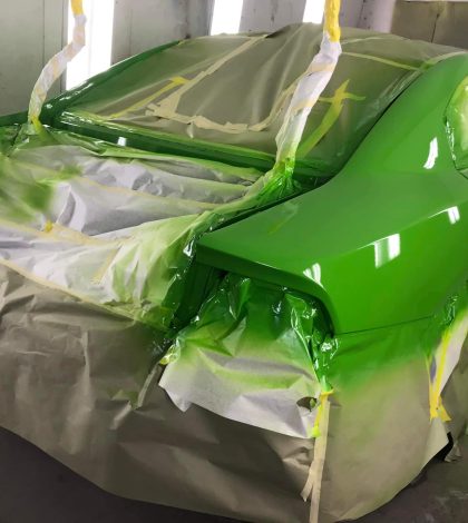 A car partially covered in green paint, masked with paper and tape, in a spray booth with bright lights.