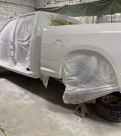 A partially painted white pickup truck in a workshop, wrapped in plastic for protection, with tools and equipment in the background.
