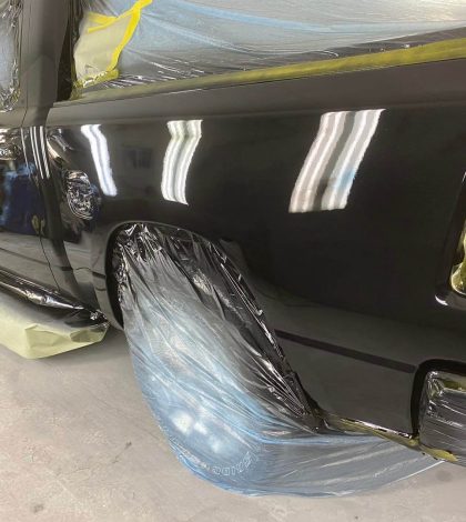 A truck is being painted inside a garage, with paint supplies and tools visible in the background.