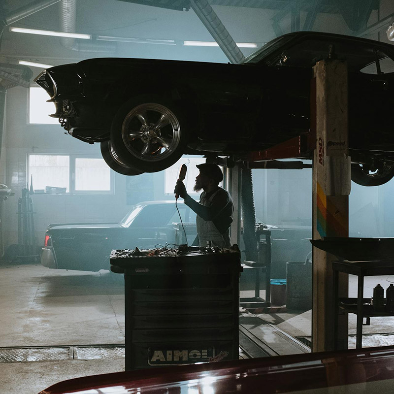 A mechanic works under a raised classic car in a dimly lit garage filled with tools and shadows.
