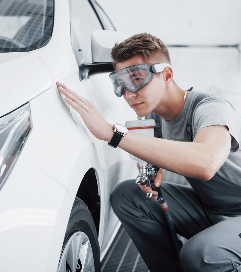 A man in blue overalls painting a car with a spray gun in a well-lit garage.